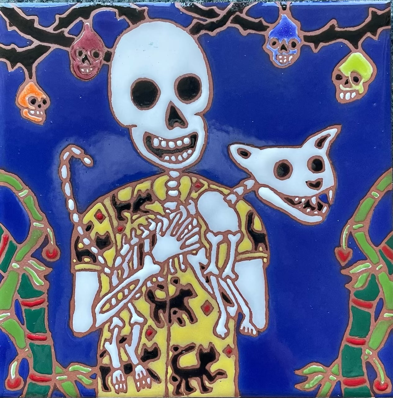 Artistic ceramic tile depicting a skeleton figure holding a cat, surrounded by vibrant colors and playful skull motifs, designed by Hearts for the Arts for the Hearts for the Arts | 2 Tiles collection.