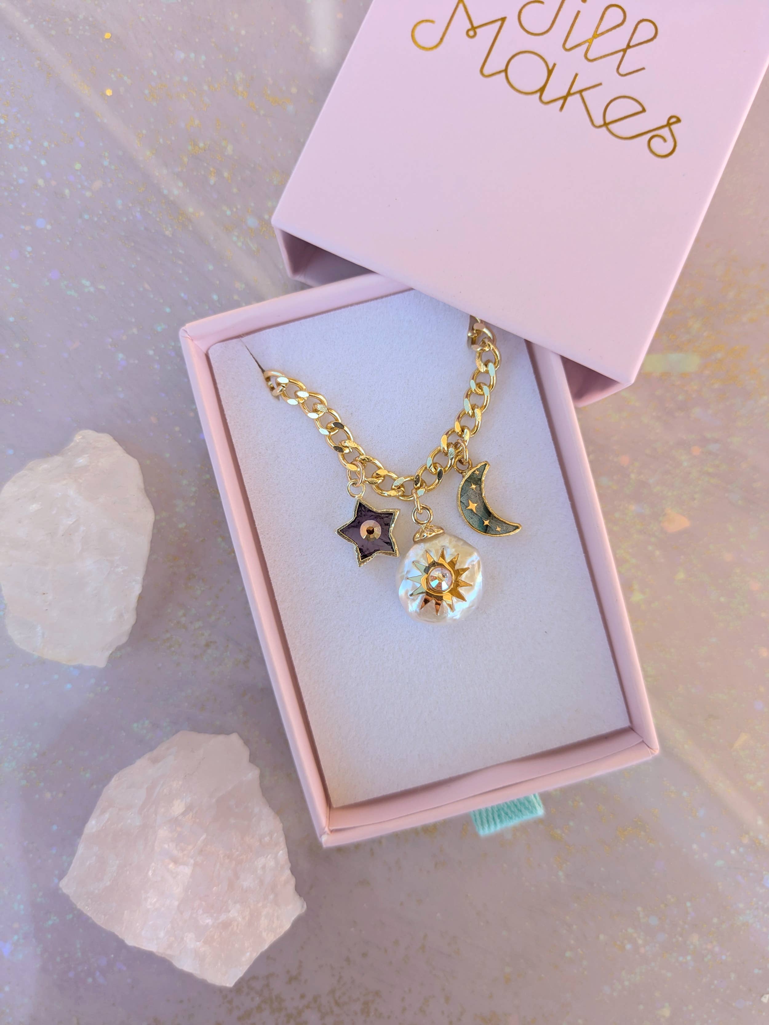Elegant presentation of the Pearl and Moon Celestial Charm Necklace by Jill Makes in a pink box, featuring gold charms of a pearl sun, amethyst star, and labradorite moon, surrounded by crystals.