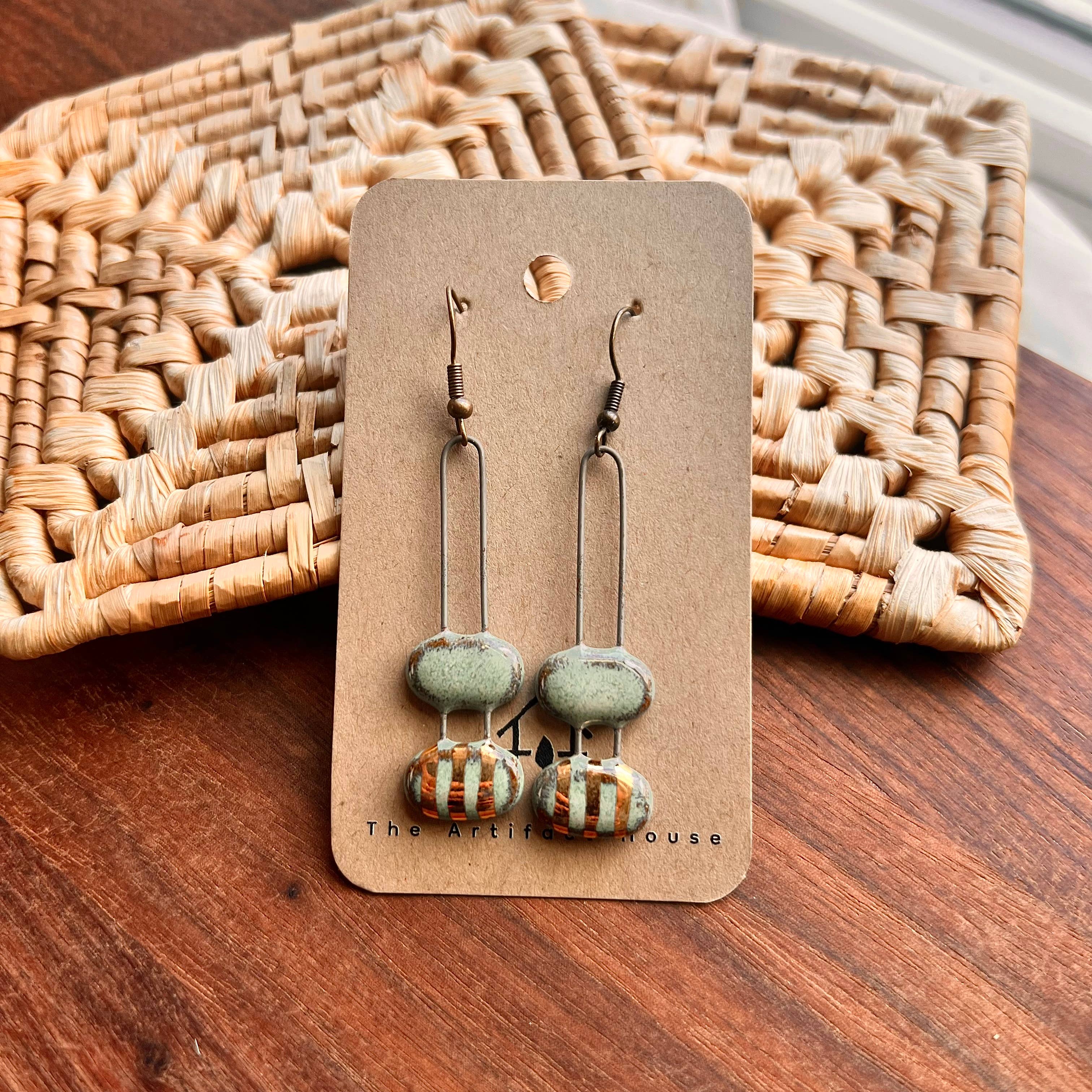 Handmade turquoise double bubble gold striped ceramic earrings displayed on a card by The Artifact House, set against a woven background.