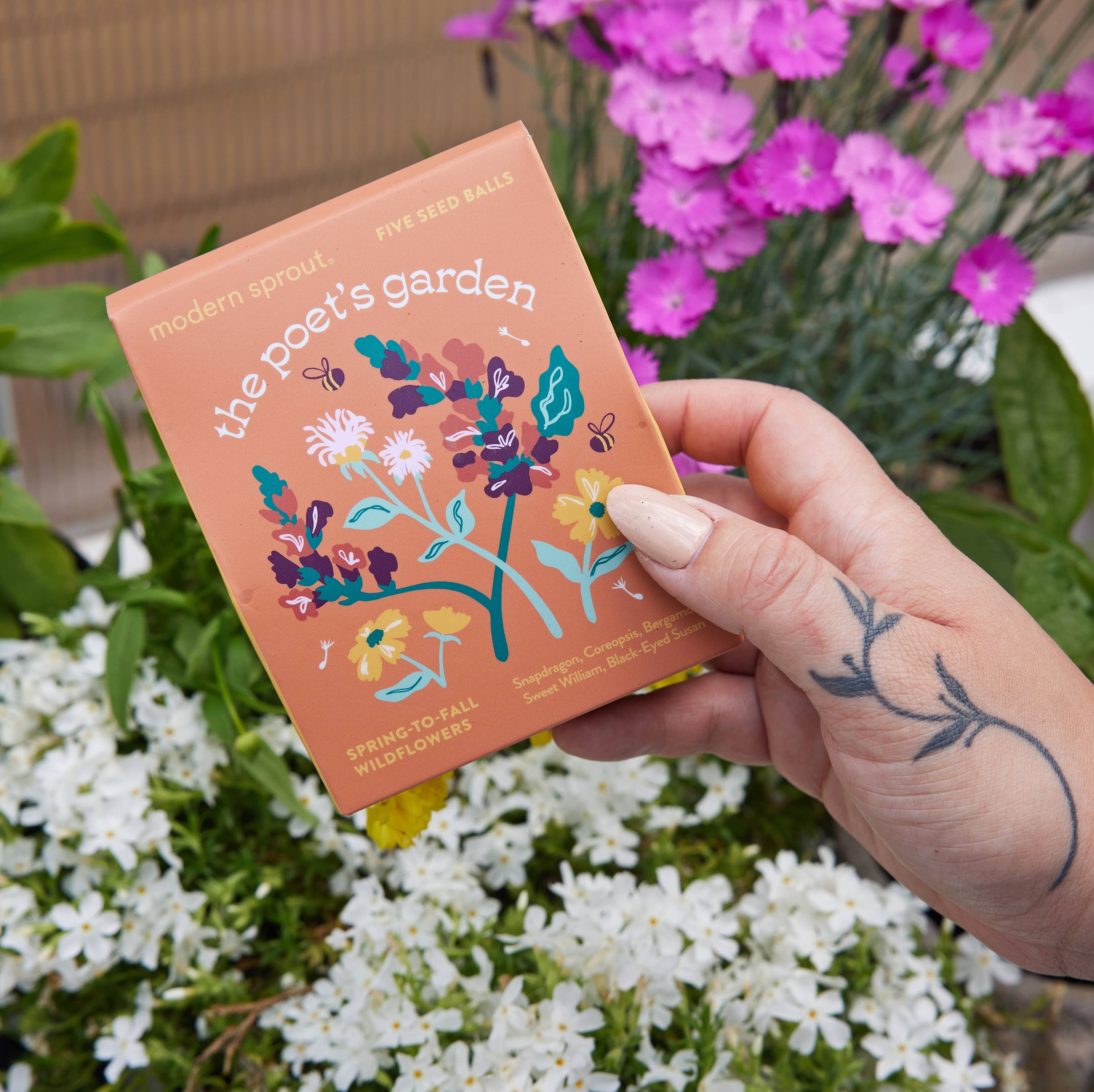 A close-up of The Poet's Garden seed ball kit from the Bespoke Blooms collection by Modern Sprout, held in hand with blooming flowers in the background.