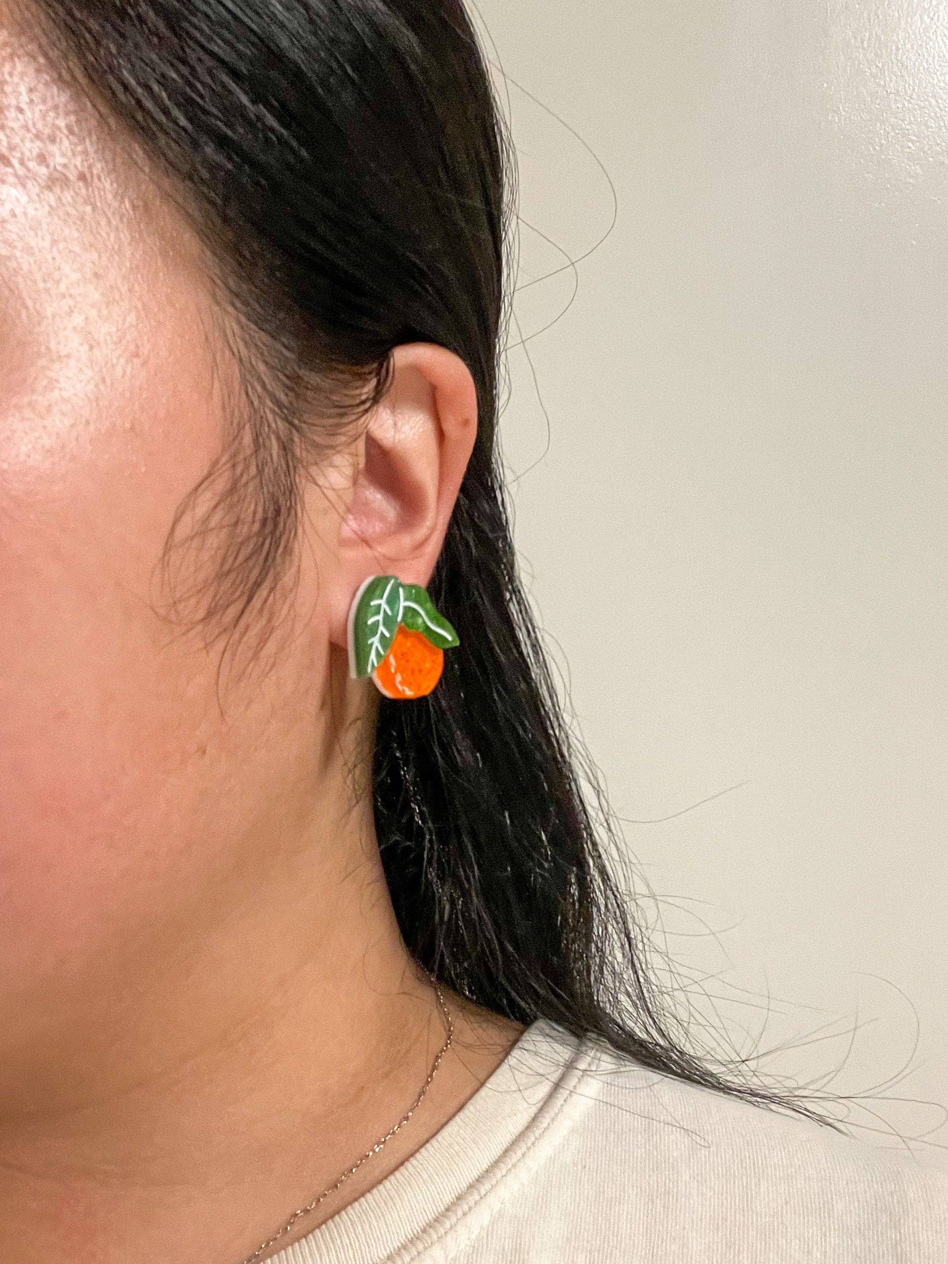Close-up of a model wearing the Mini Orange Dangle earrings from Sunnie Creative, featuring orange acrylic fruit design and green leaves, highlighting its unique handmade quality.
