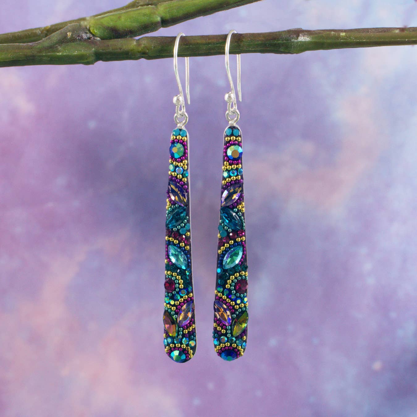 Crystal Dash Earrings by Mosaico Jewels displayed on a branch, showcasing their colorful handcrafted design and elongated silhouette against a soft purple background.