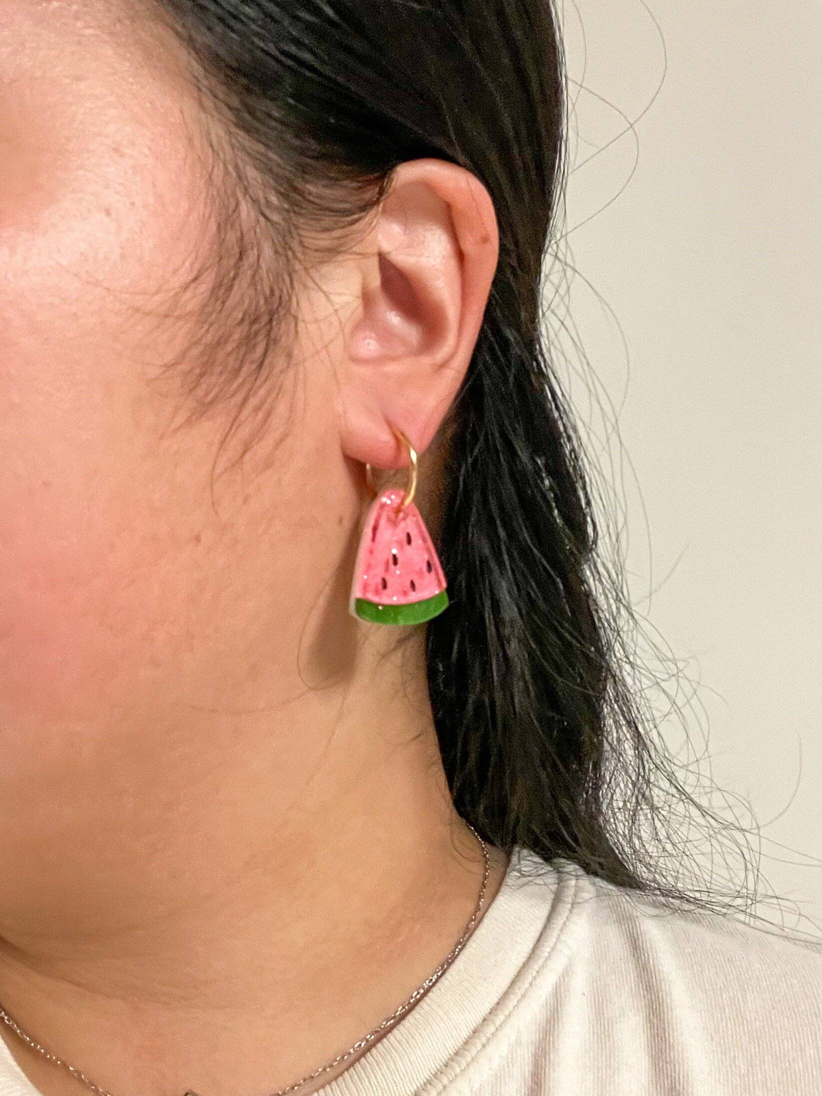 Close-up view of a person wearing Mini Watermelon Dangle earrings by Sunnie Creative, emphasizing the vibrant pink and green colors of the handmade earrings against a neutral background.