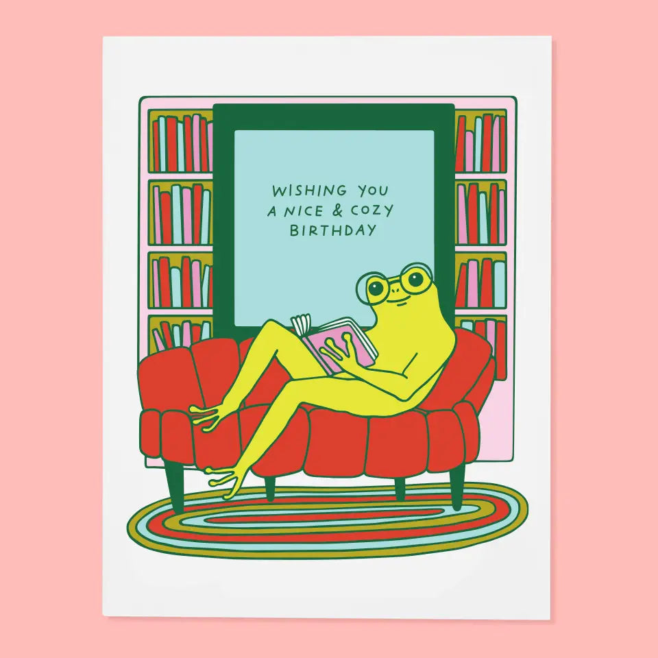 Cozy birthday card featuring a frog relaxing on a red couch with a book, accompanied by the text 'WISHING YOU A NICE & COZY BIRTHDAY', part of The Good Twin's 'The Good Twin Card Collection'.