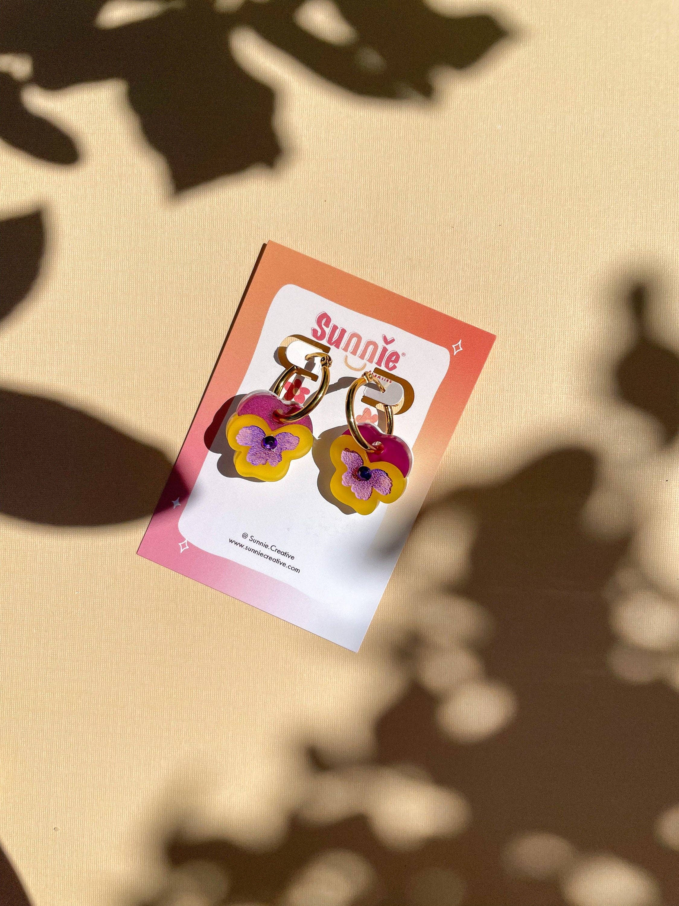 Mini Yellow Pansy Dangle earrings from Sunnie Creative displayed on a promotional card, featuring unique handmade acrylic and stainless steel design.