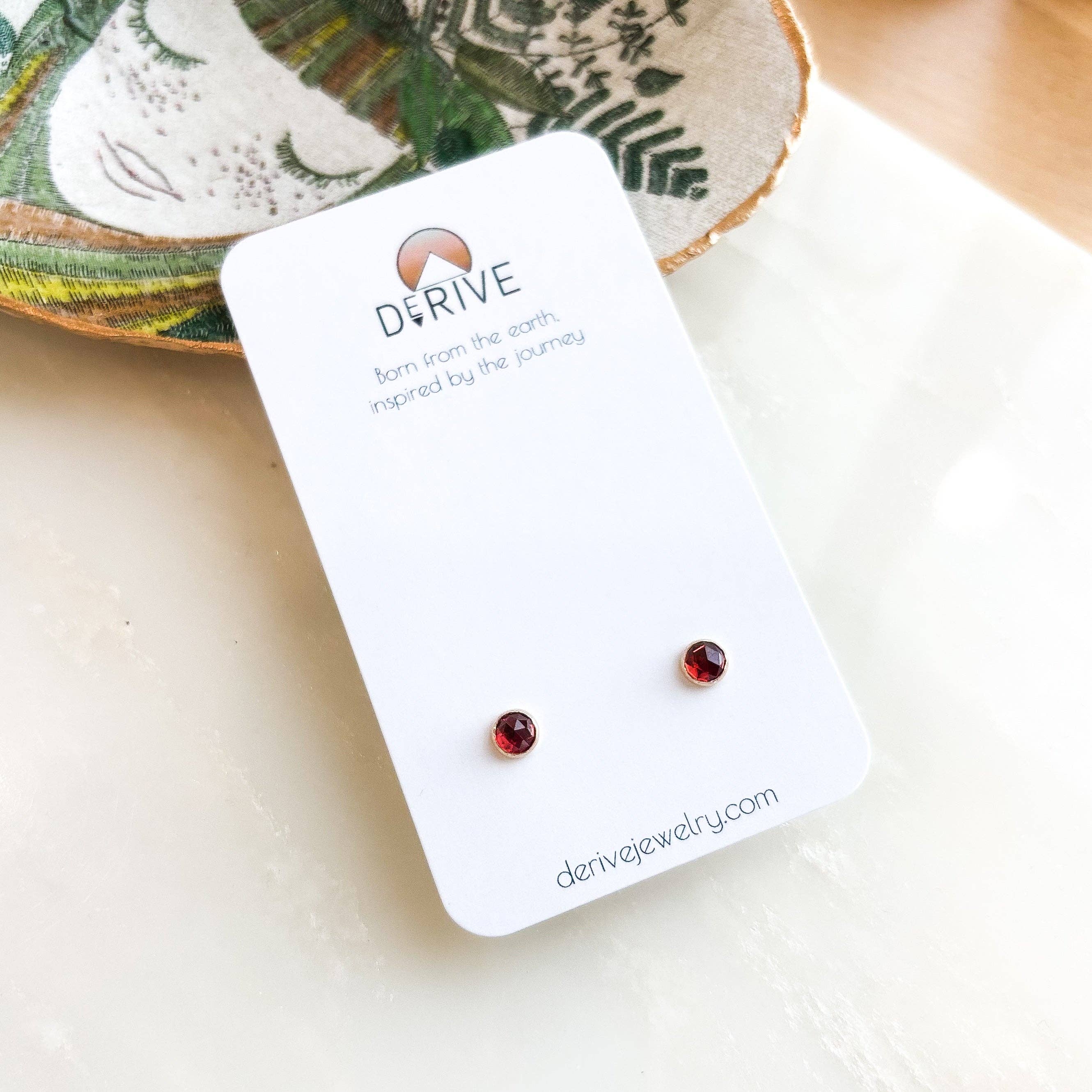 Gemstone Stud Earrings by Derive Jewelry featuring a close-up view of garnet stones set in gold fill on a display card.