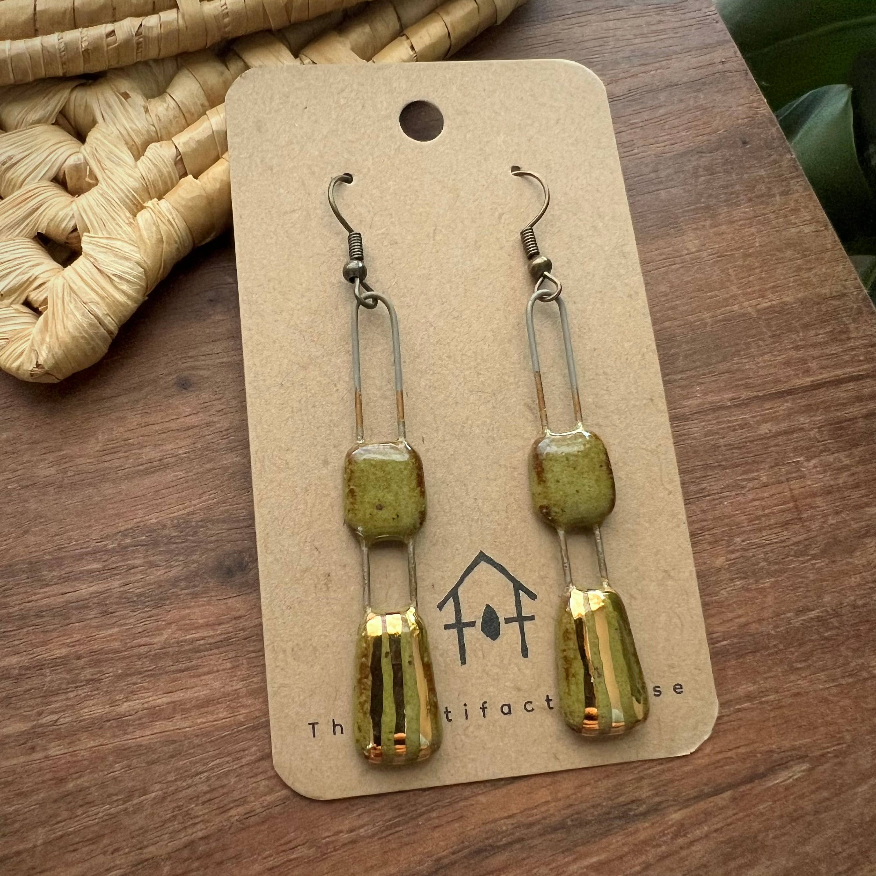 Speckled Olive Two Piece Gold Lined Earrings displayed on a card by The Artifact House, set against a wooden surface and woven basket.