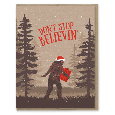 Humorous Christmas card showcasing a Sasquatch in a Santa hat carrying presents with the phrase 'Don't Stop Believin'' by ARTISANS & agency from the Christmas Card Collection.