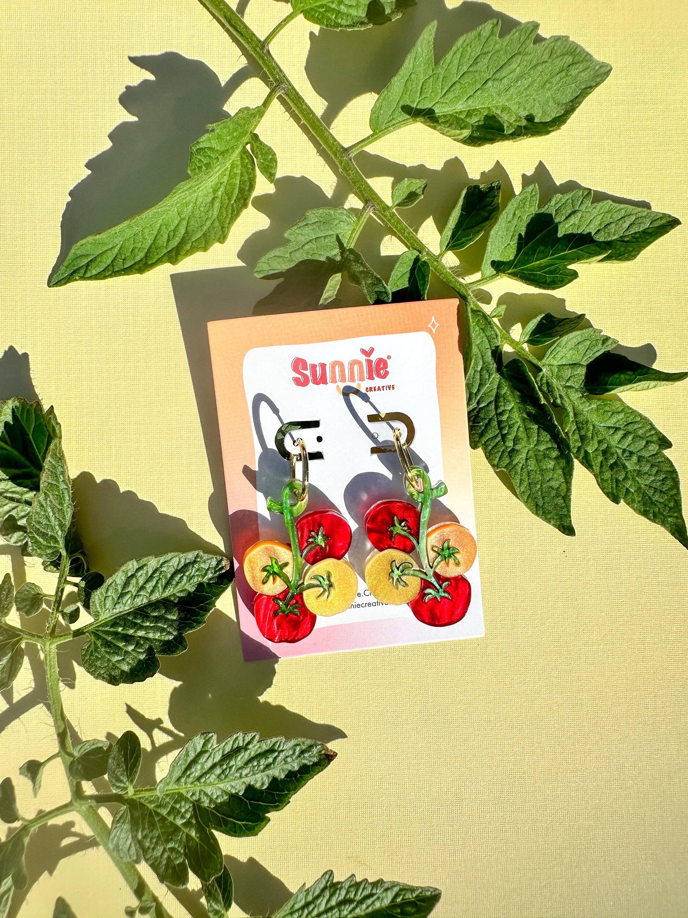 Stylish display of Cherry Tomato Dangle earrings by Sunnie Creative, presented on a card with a backdrop of fresh tomato leaves, emphasizing the colorful cherry tomato design.