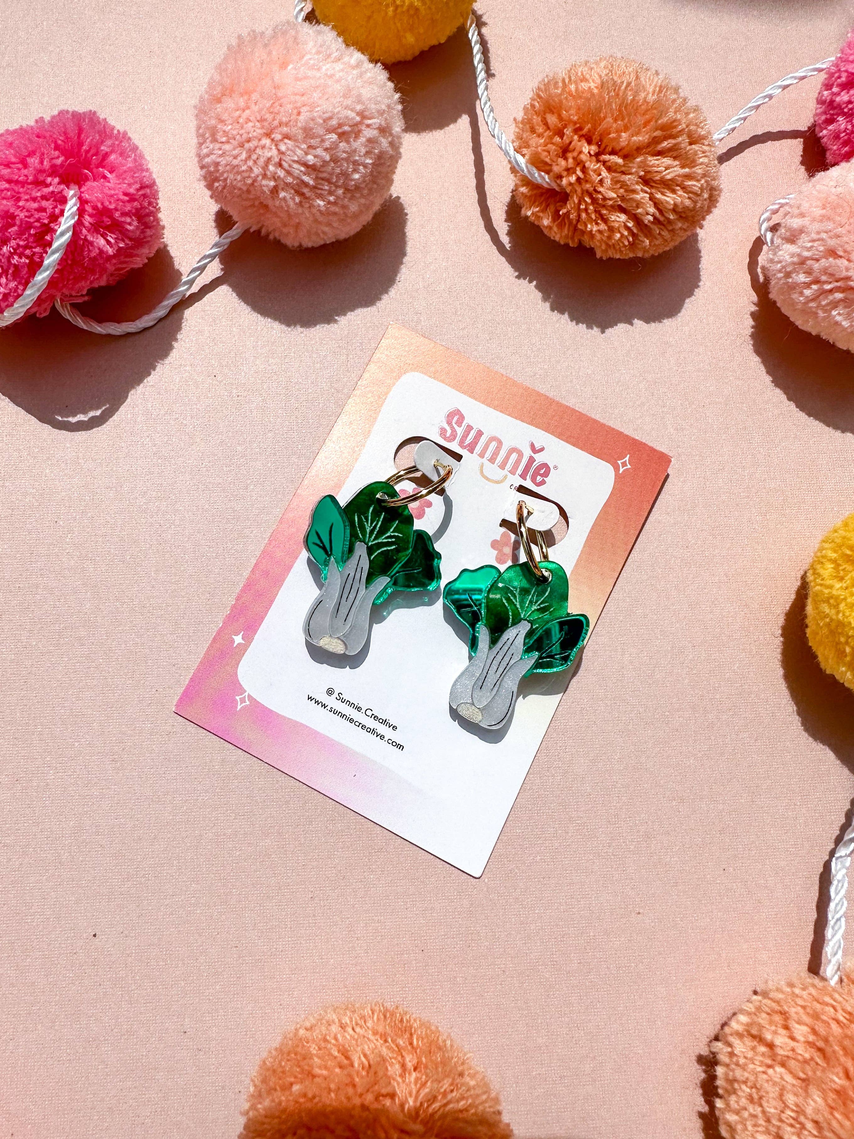 Bok Choy Dangle earrings by Sunnie Creative presented on a card surrounded by colorful pom poms, emphasizing their playful and artistic style.