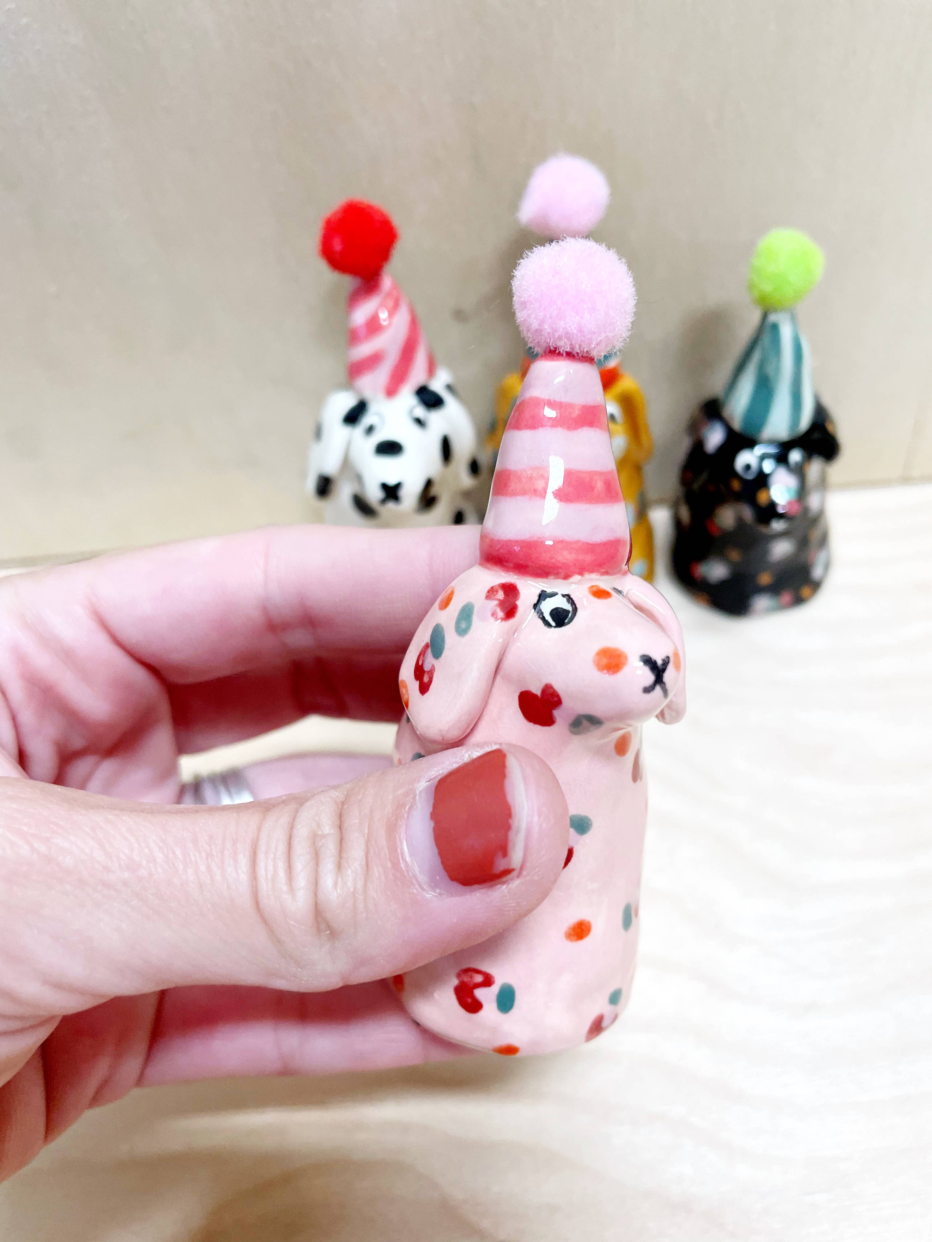 Lovely mini party dog in soft pink with colorful dots and a striped red party hat, created by Bianca Rae. A delightful piece from 'Party Minis! Mini Party Dogs in Party Hats.'