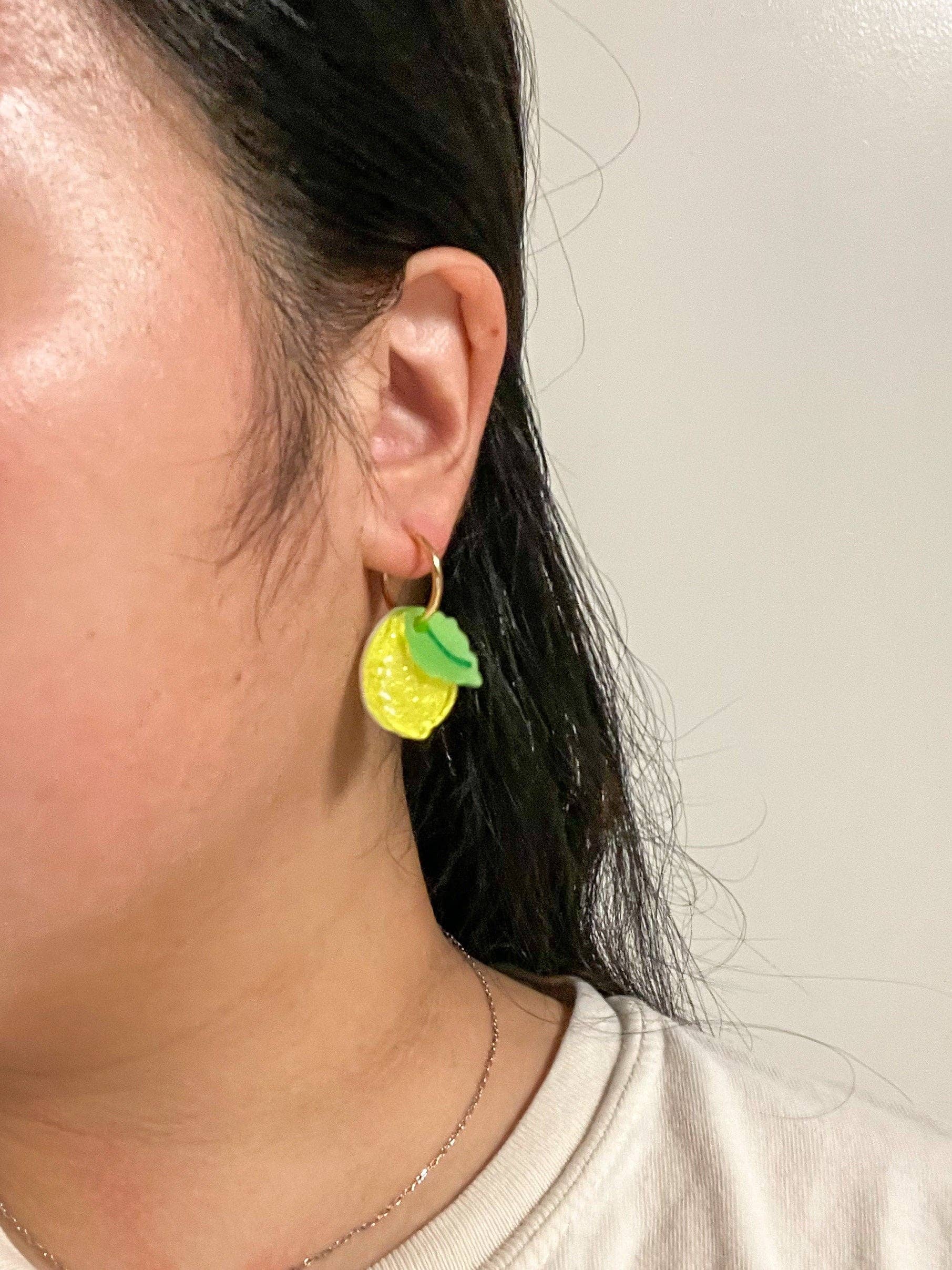 A person wearing Mini Lemon Dangle earrings, highlighting the unique yellow acrylic design with green leaf accents made by Sunnie Creative.