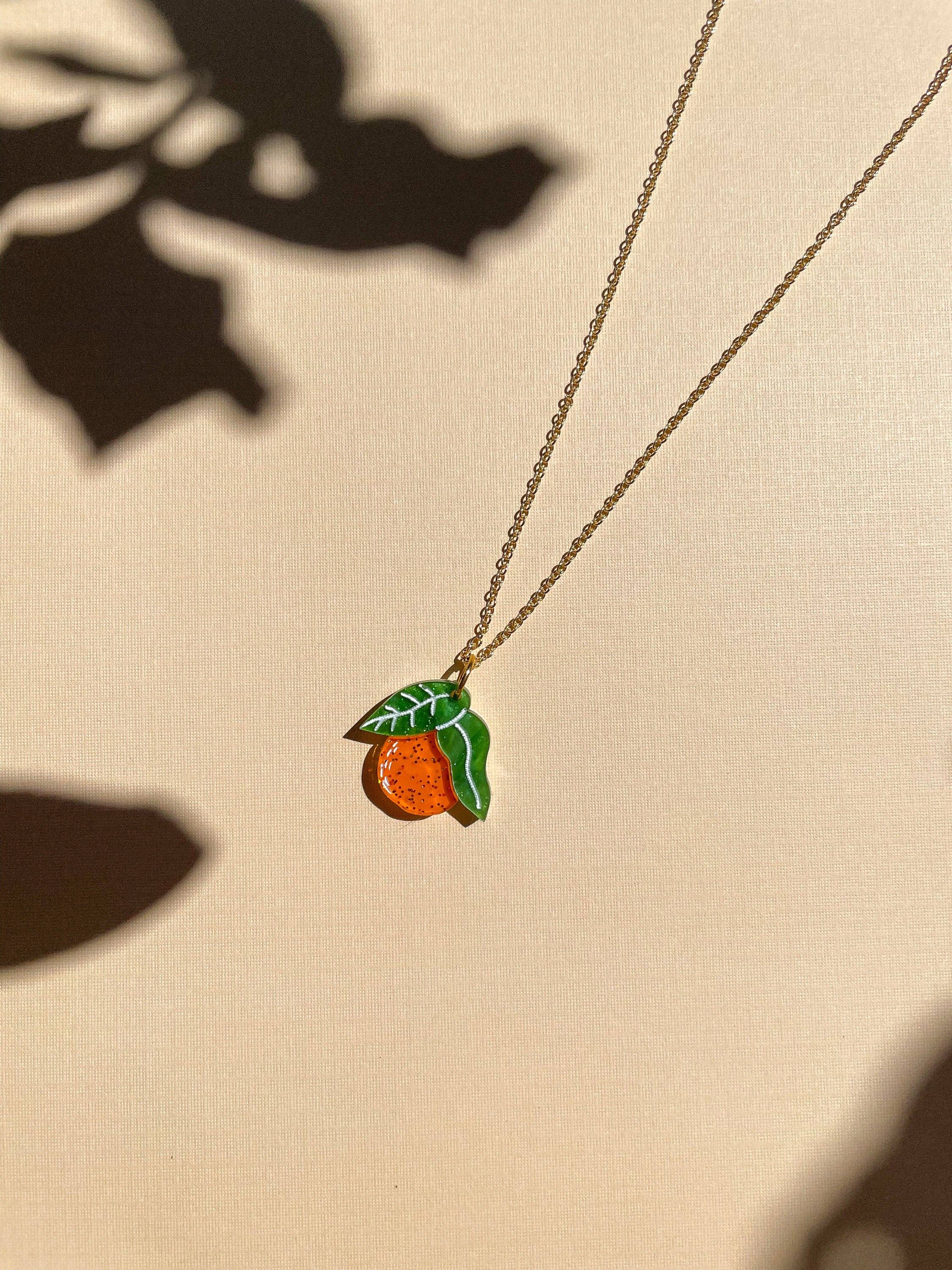 Mini Orange Necklace by Sunnie Creative, featuring a vibrant orange pendant with green leaves, resting on a soft background, showcasing the unique handmade design.