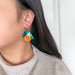 Close-up of a model wearing Orange Dangle earrings by Sunnie Creative, showcasing vibrant colors and stylish design against a neutral backdrop.