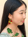 A model wearing Cherry Tomato Dangle earrings by Sunnie Creative, highlighting the playful cherry tomato design against her long black hair, with green foliage in the foreground.