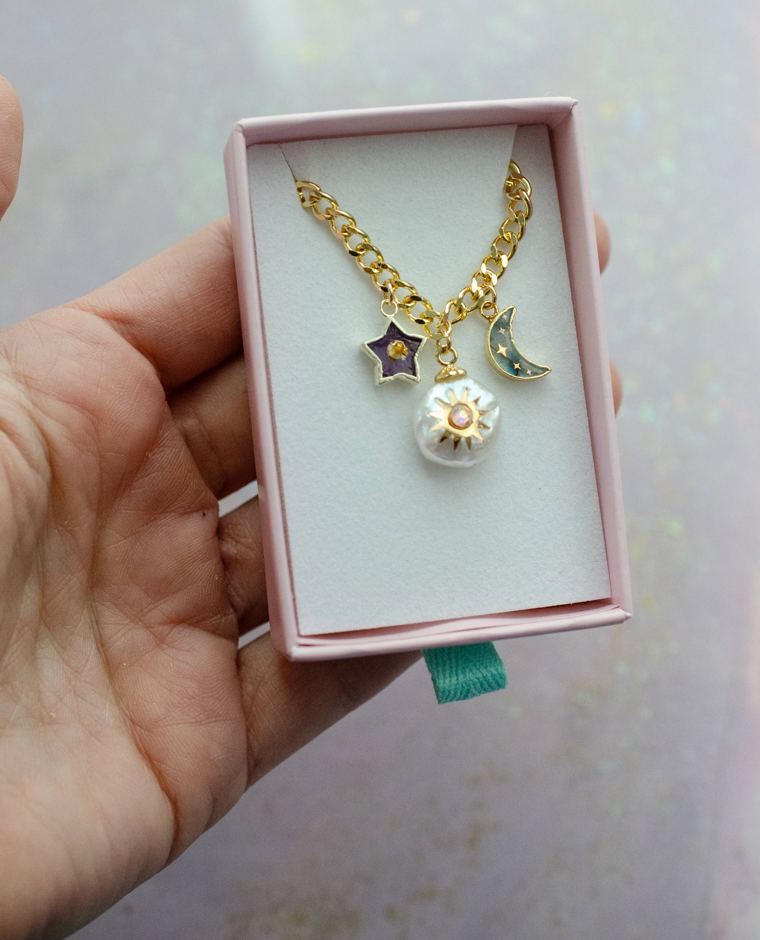 Hand holding a gold curb chain necklace with celestial charms, including an amethyst star, a pearl sun, and a labradorite moon, packaged by Jill Makes, titled 'Pearl and Moon Celestial Charm Necklace'.