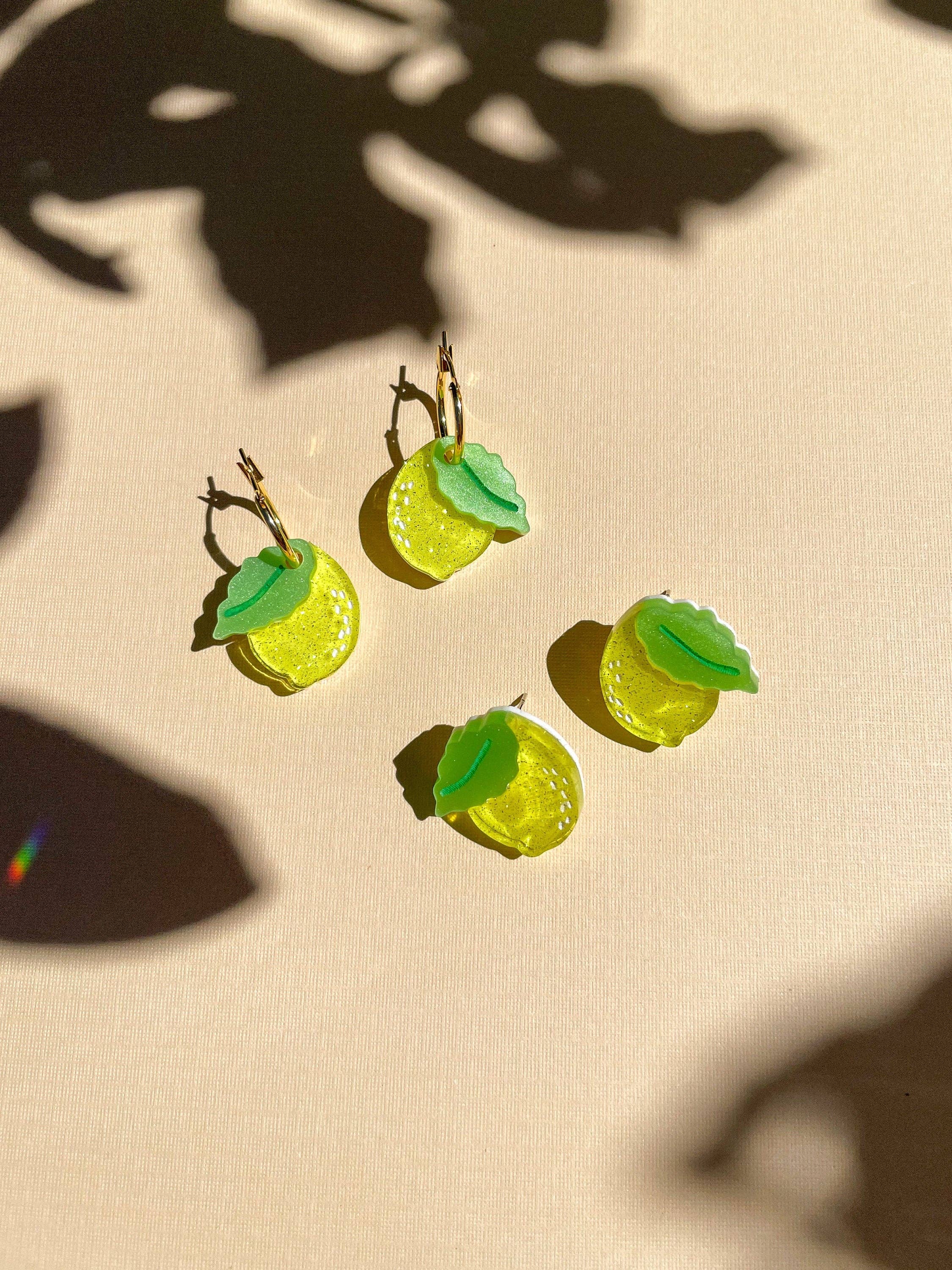 Mini Lemon Dangle earrings displayed on a textured background with shadow play, featuring vibrant yellow acrylic with green leaf accents by Sunnie Creative.