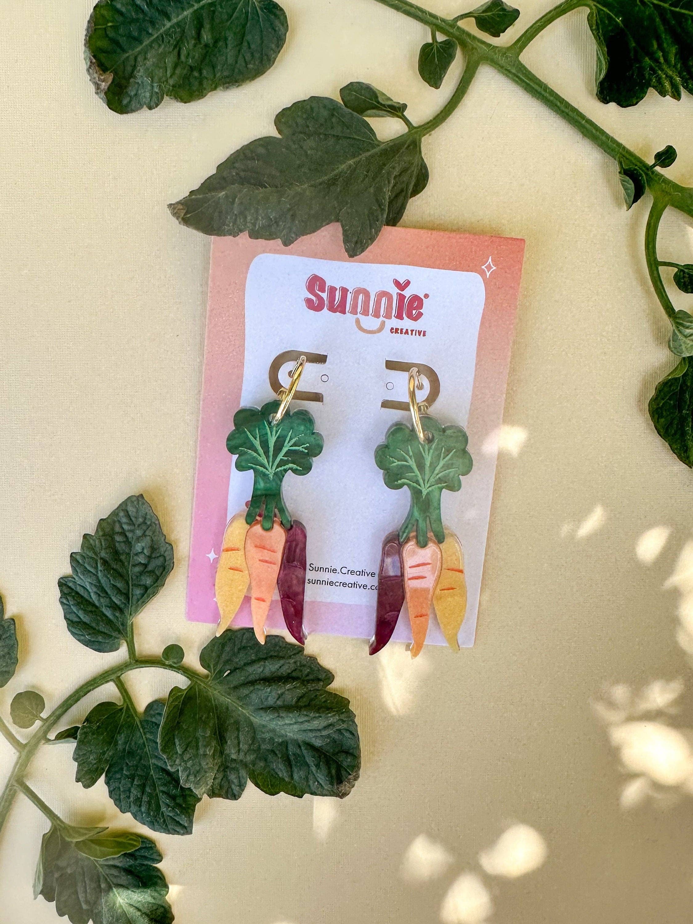 Close-up of Rainbow Carrot Dangle earrings by Sunnie Creative displayed on a card, featuring unique carrot designs made of acrylic with gold-plated brass hoops.