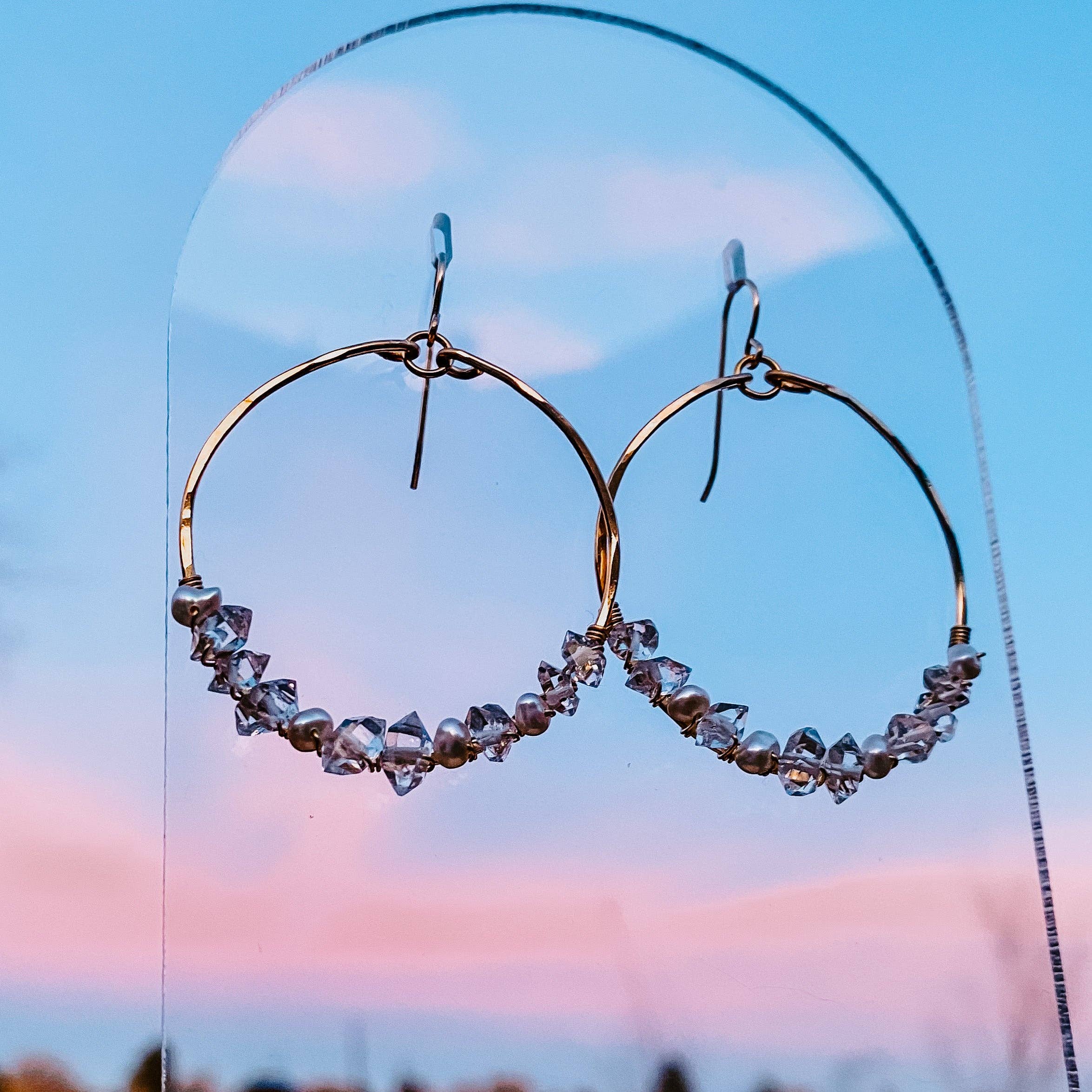 Artistic shot of Everly Herkimer Diamond and Pearl Hoop Earrings by Derive Jewelry against a sunset background, featuring crystal-like diamonds and luminous pearls on a gold hoop.