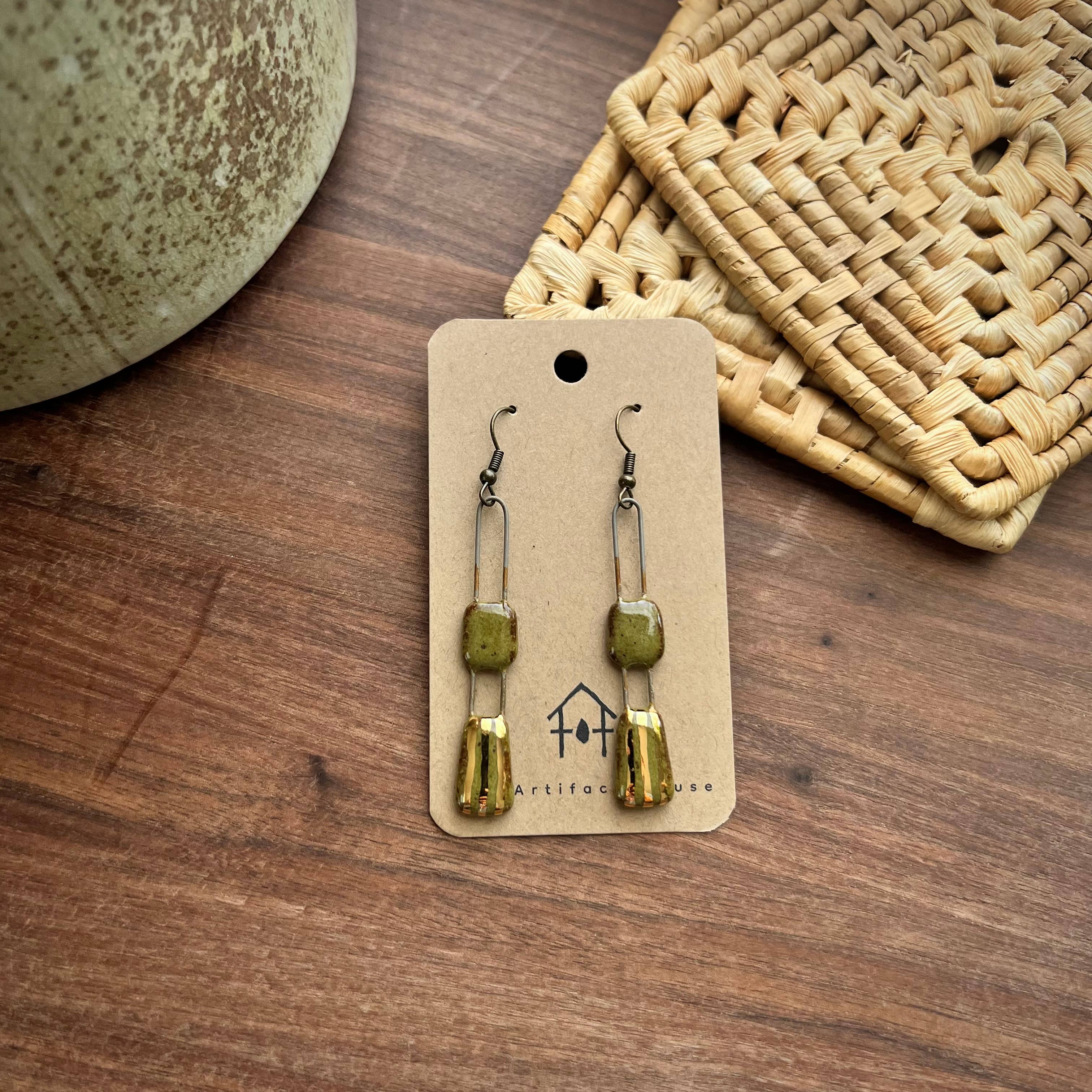 Single pair of Speckled Olive Two Piece Gold Lined Earrings on a card by The Artifact House, resting on a wooden surface with a woven basket.