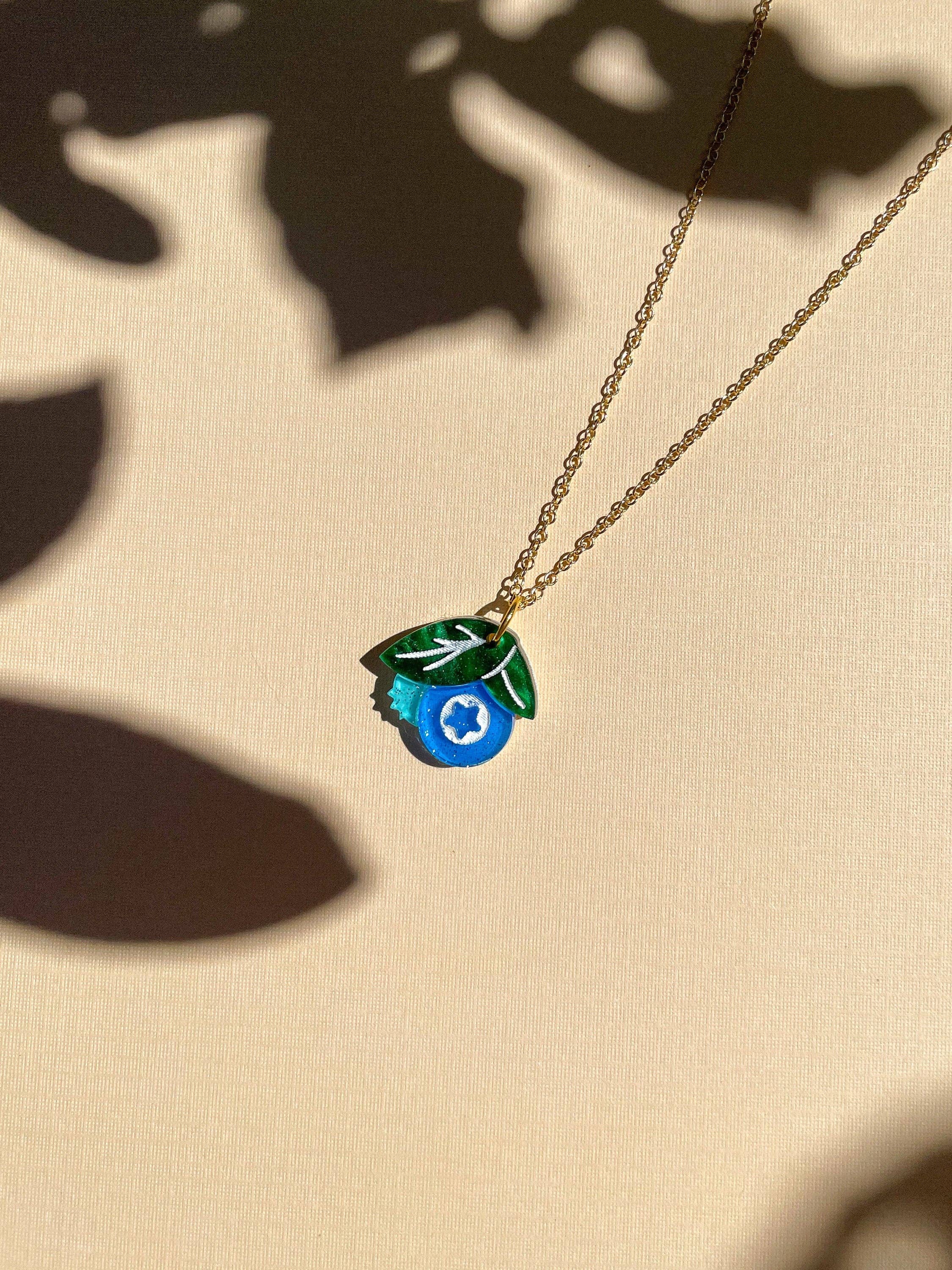 Mini Blueberry Necklace by Sunnie Creative featuring a vibrant acrylic blueberry pendant with green leaf, showcased in soft lighting against a textured background.