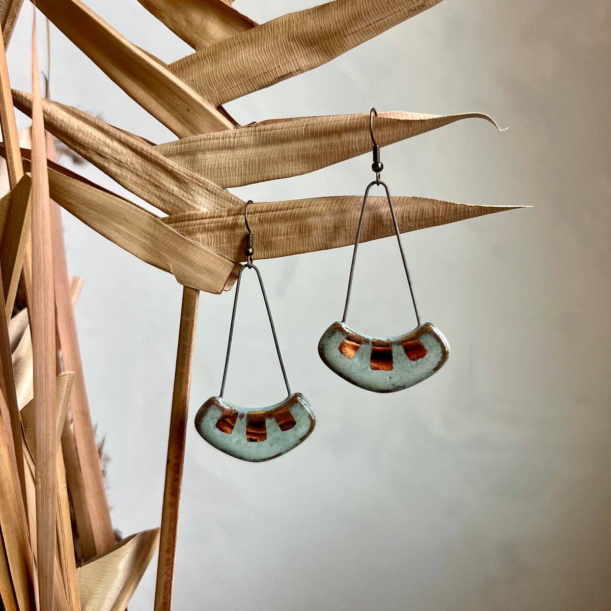 Turquoise and Gold Collar Earrings by The Artifact House, elegantly hanging among dried plant leaves, highlighting the unique ceramic craftsmanship and artistic touch.