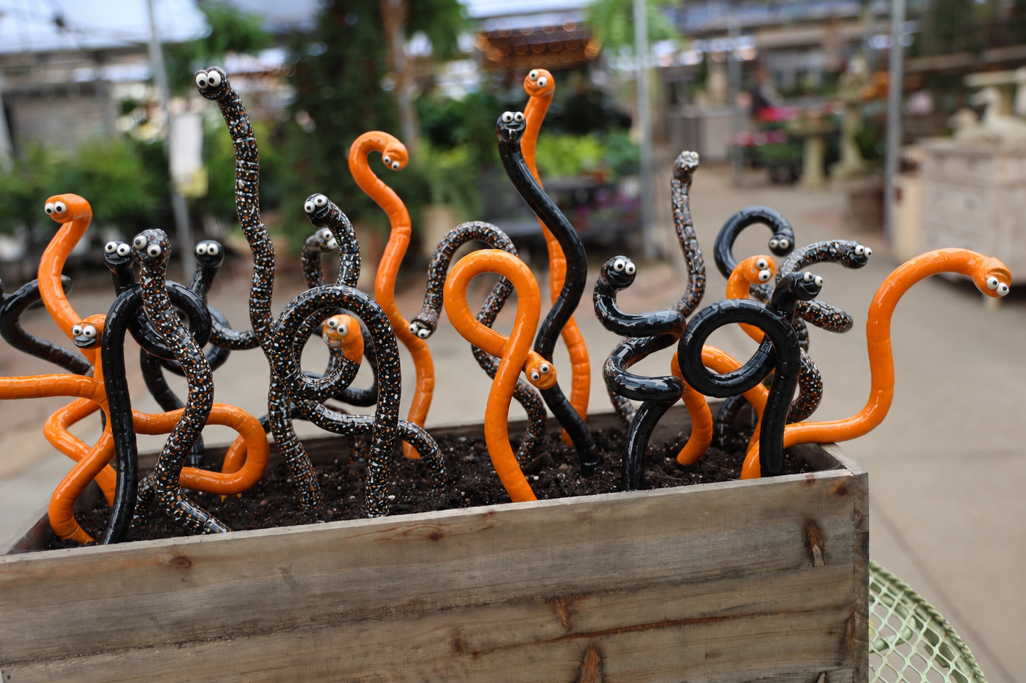 A wooden planter box filled with multiple curled, snake-like sculptures in glossy black and bright orange, photographed outdoors with a blurred garden or market background.